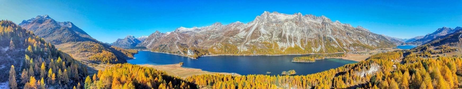 Bergpanorama im goldenen Herbst bei Sonnenschein und blauen Himmel. Im Vordergrund einige Bäume, ein See und im Hintergrund eine Bergkette.