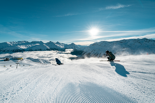 Zwei Skifahrer die eine Piste bei blauen Himmel herunter fahren. Die Sonne steht hoch am Himmel und der Himmel ist frei von Wolken