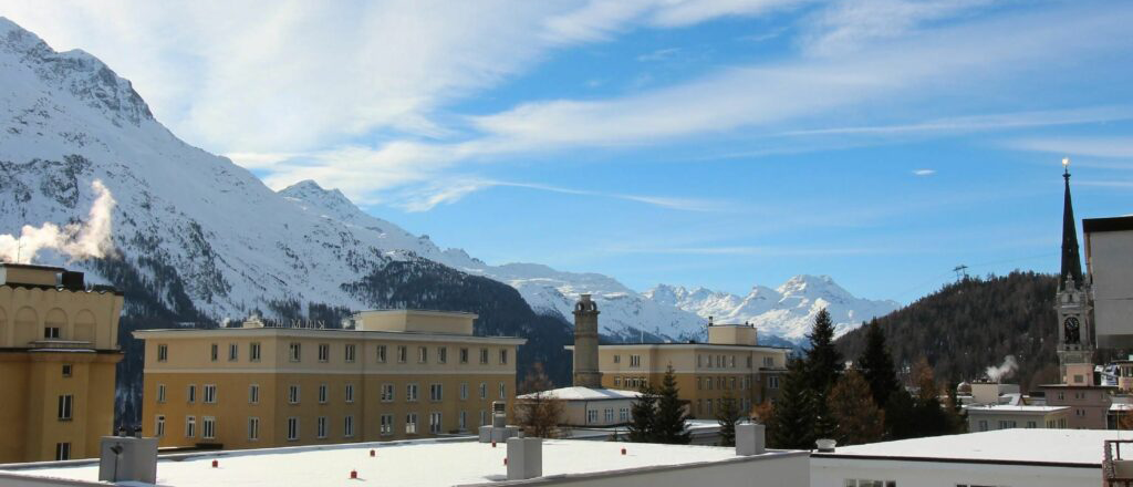 Über den Dächern der Immobilien im Engadin. Man erkennt die Bergkette, Sonnenschein und einen blauen Himmel.