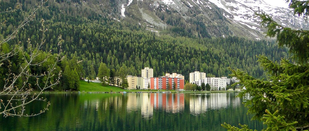 Ein See mit einer Hochhausreihe am Ufer mit einer Bergkette und einem Waldstück dahinter, im Sommer.