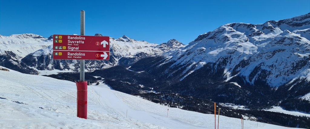 Ein Pass auf einem Berg mitten im Winter. Mittig erkennt man einen roten Wegweiser und im Hintergrund eine schneebedeckte Bergkette.